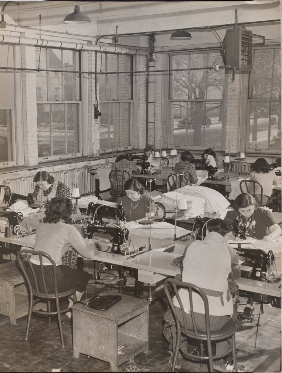 Women Seated at Desks Work on Sewing Machines by Arnold Eagle