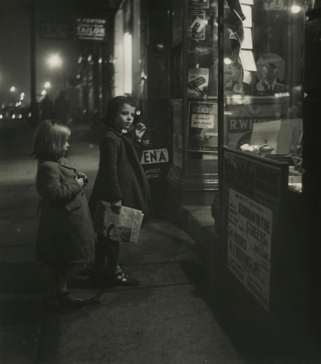 Two Little Girls in Winter Coats Standing Outside a Shop at Night by Godfrey Macdominic