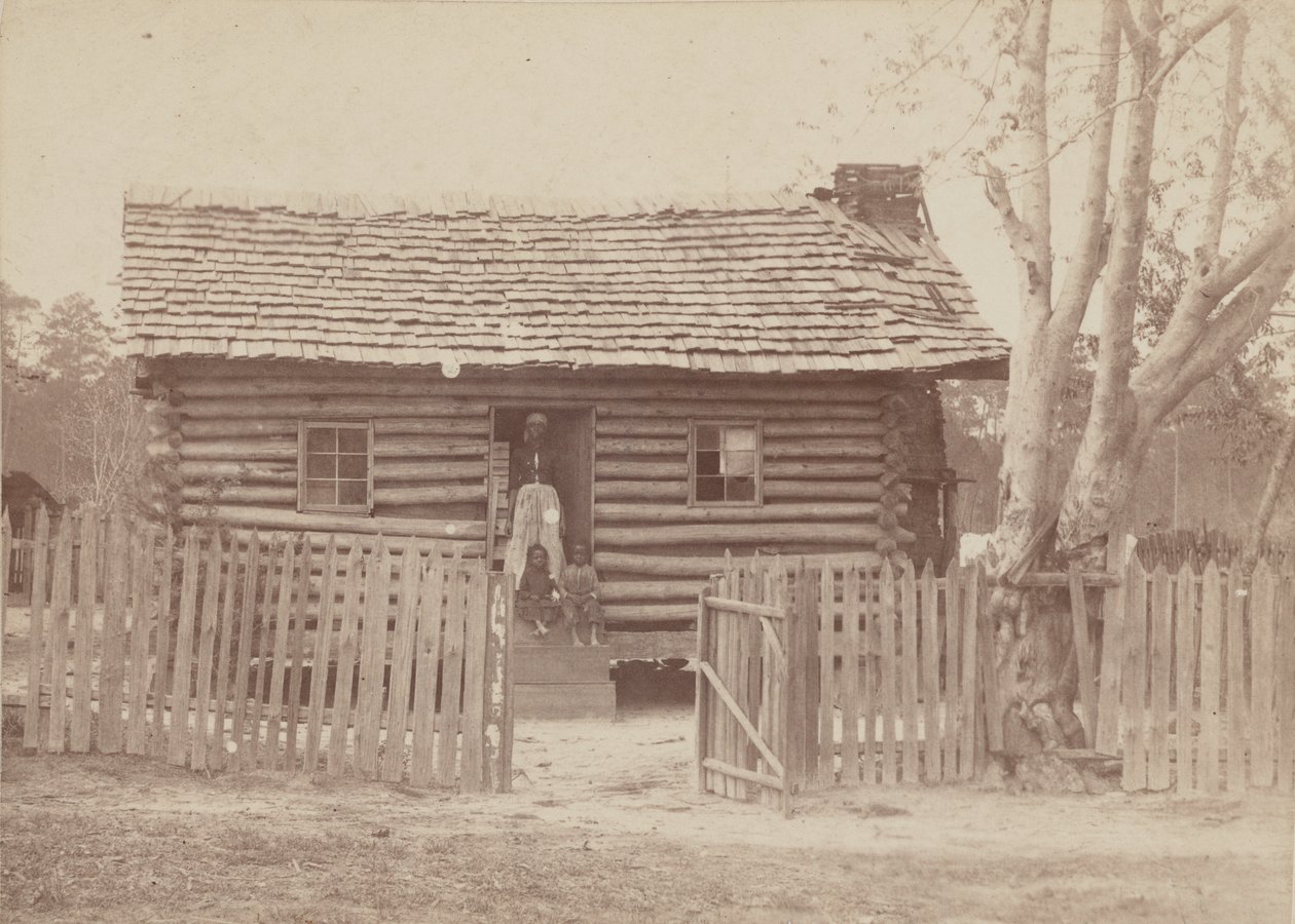 Albumen print of a woman and two children in front of a log house in Georgia by H. S. Clark