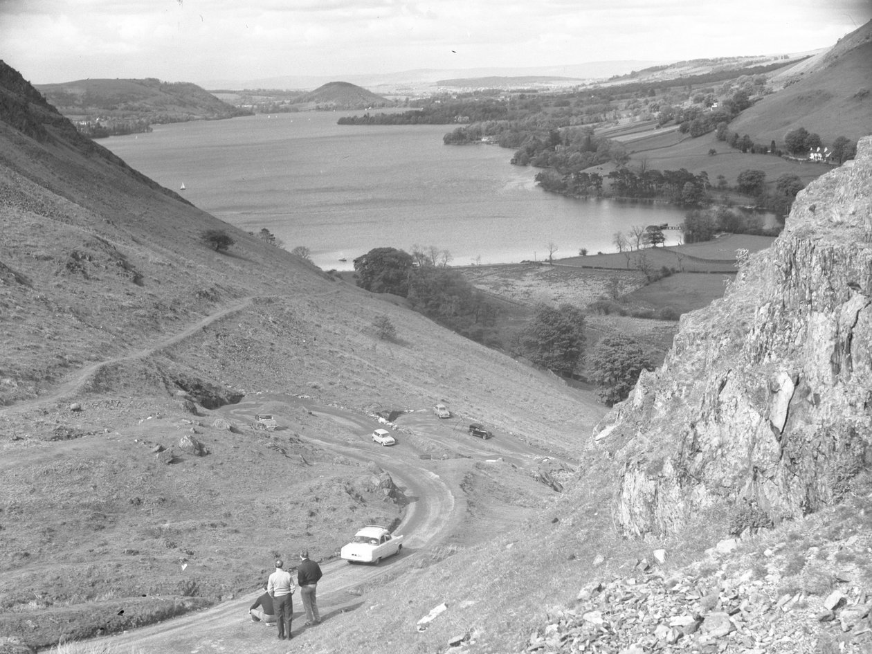 A view looking down a fellside with road, cars and people on, lake in background by Joseph Hardman
