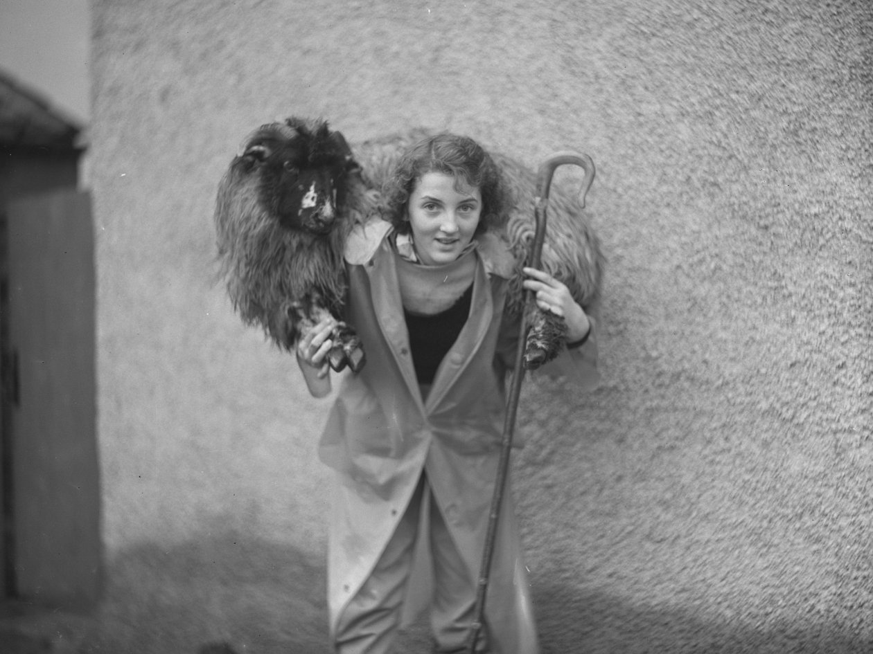 A woman stands with a sheep across her shoulders while holding a shepherd