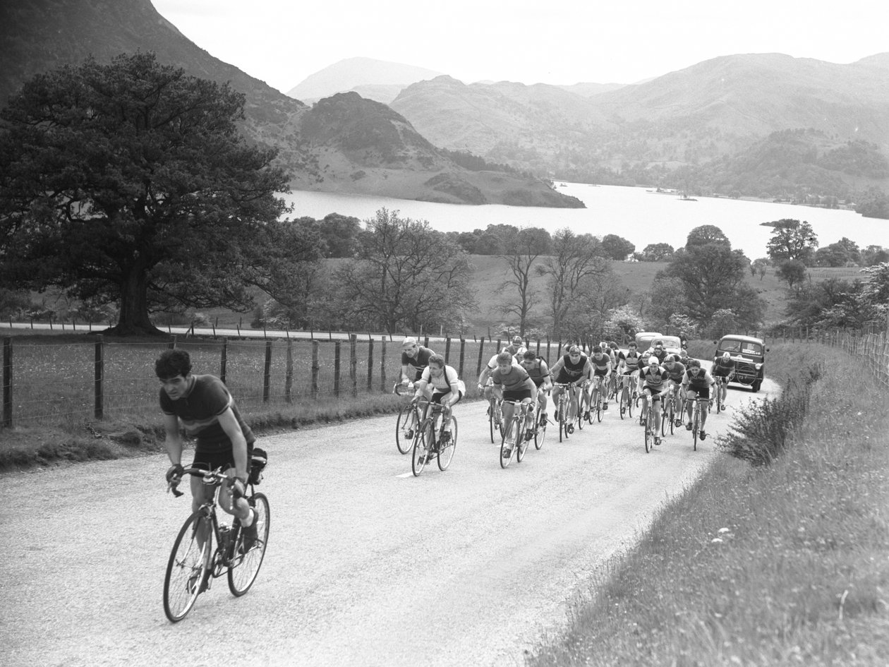 Group of cyclists heading up a road followed by cars, lake and hills in background by Joseph Hardman