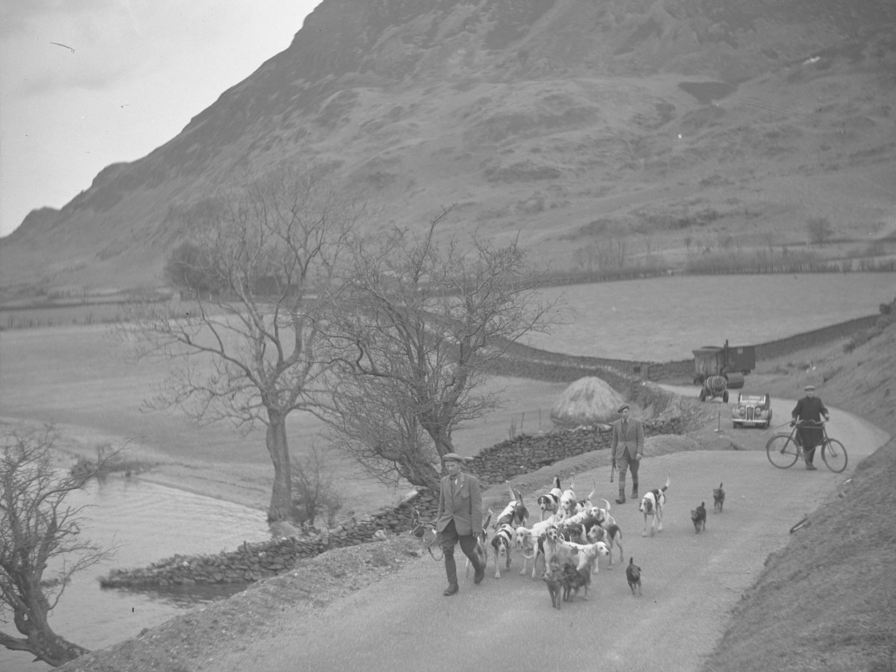Two men walking with hounds in a lane, man with a bike and machinery, haystack and lake to the left, hills in background by Joseph Hardman