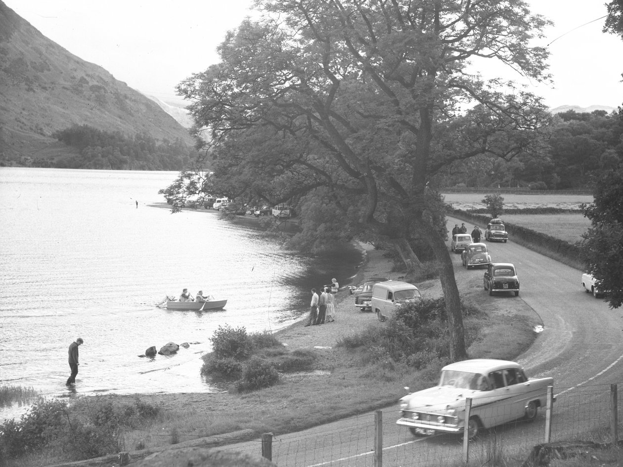 View of lake shore with people standing, walking, wading and boating, road runs to right of image with parked cars by Joseph Hardman