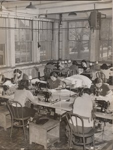 Women Seated at Desks Work on Sewing Machines by Arnold Eagle