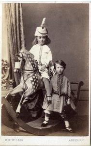 Young girl with hat on rocking horse with another girl standing by
