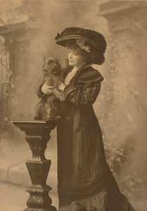 Sarah Bernhardt Standing in a Studio Setting, Holding a Little Black Dog on an Ornate, Carved Wooden Pedestal