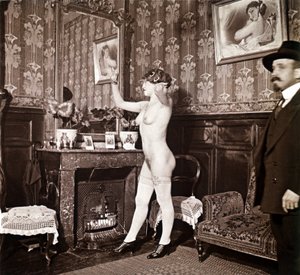 Enigmatic photograph showing a young woman completely naked (except for her shoes and stockings) recoiffing her hair in front of a mirror in the presence of a man dressed and heated with a hat. One might think it was a photo taken in a brothel, but this hy by Unbekannter Künstler