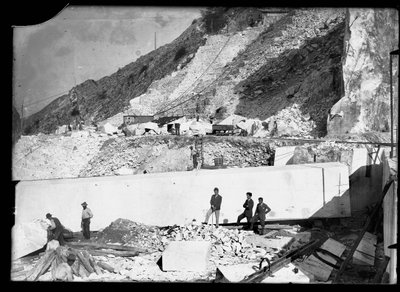 Three men observe the work of several excavators in the Apuan Alps. Behind them is an enormous block of marble already squared off and ready to be transported to the valley by A. Corsini