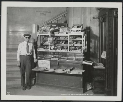 Blind news dealer, John Martie, at his stand at the south end of the Municipal Building, New York by Company Byron
