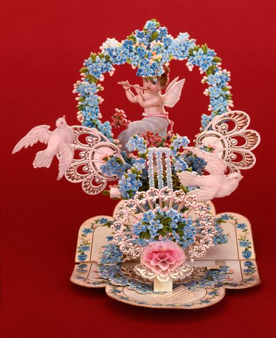 A pop-up card a winged cherub playing a flute under a heart shaped arch of forget-me-nots by English School
