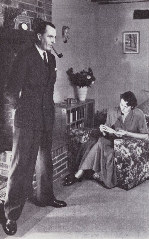 Wife reads, while husband warms his back against the fire by English Photographer