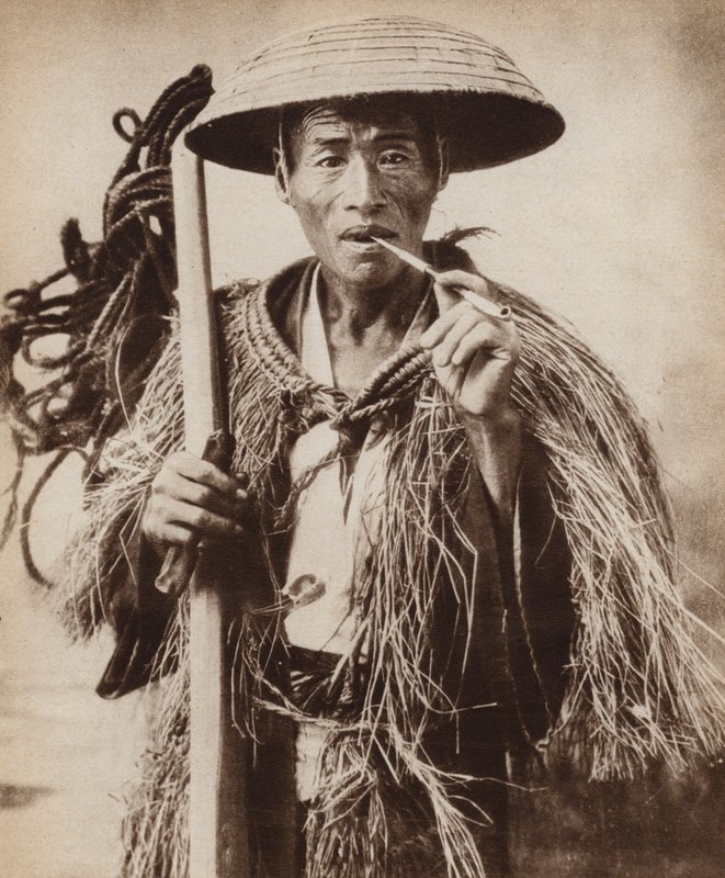 Japanese coolie (farm labourer) wearing a hat to protect him from heavy rain in the rice fields by Photographer Japanese