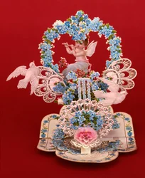 A pop-up card a winged cherub playing a flute under a heart shaped arch of forget-me-nots