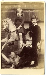 Young girl on rocking horse with one boy standing and one boy seated with dog
