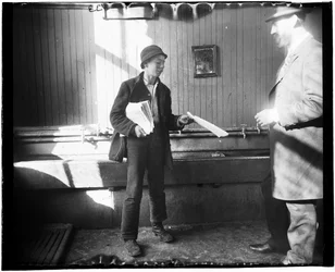 Portrait of a Newsboy Handing a Paper to a Man in What Appears to Be a Restroom at the Duane Street Lodging House