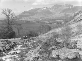 Walkers crossing a dry stone bridge over a stream, hills and lake in background