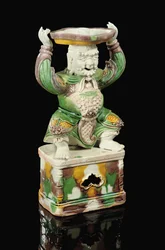 Egg and spinach glazed pricket-stick, modelled as a smiling immortal with scrolling beard, arms raised to support the quatrefoil pricket attachment balanced on his head, Kangxi