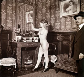 Enigmatic photograph showing a young woman completely naked (except for her shoes and stockings) recoiffing her hair in front of a mirror in the presence of a man dressed and wearing a hat. One might think it was a photo taken in a brothel, but this hyp