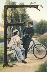 F: A Man in Cyclist Outfit Leaning on His Bicycle Talking to a Woman Sitting on a Bench. Countryside Setting - Alphabet Characters Ref Th. E., L. Theochrom - Series 994. Theodor Eisman, publisher, Leipzig