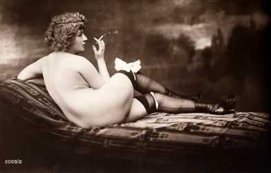 Portrait of a young woman posing naked, with her back turned, as she smokes a cigarette