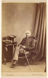 Unidentified White-Haired Man Seated, Holding a Cane, Top Hat and Book on Table at Left