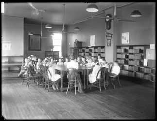 Children listen to a woman in the reading room of an unidentified branch of the Queens Borough Public Library, c.1910