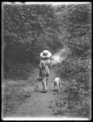 William Gray Hassler in Straw Hat and Overalls, Carrying Fishing Pole and Basket, Walking Down a Path in the Woods with a Small Dog (Bessie), August 20, 1911