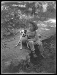William Gray Hassler in straw hat and overalls, posed seated in the woods with a small dog (Bessie)